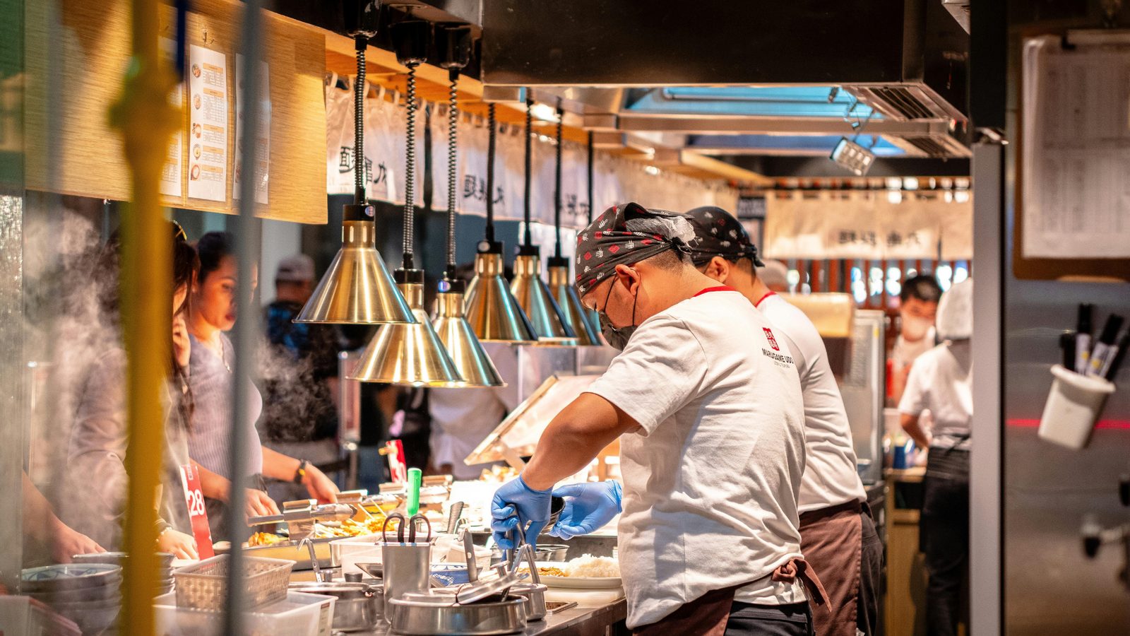 A busy restaurant kitchen line with printed menus on the wall that an assistant can read. ไลน์ครัวร้านอาหารที่คึกคักพร้อมเมนูติดผนังที่ผู้ช่วยอ่านได้